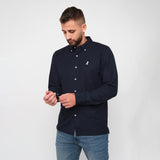 A man with a beard standing, wearing a navy blue button-down shirt, adjusting his sleeve, paired with jeans.