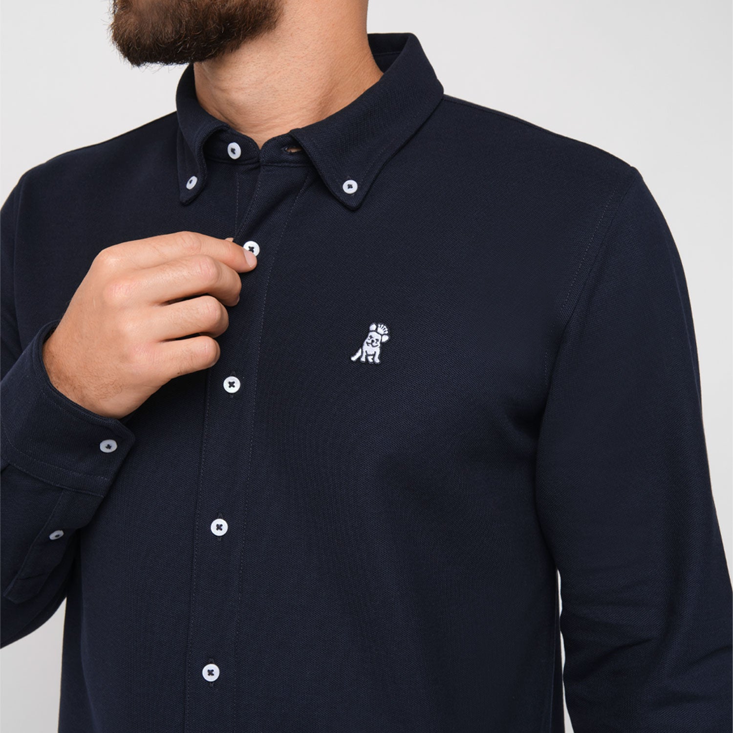 Close-up of a man wearing the navy blue button-down shirt, showing the embroidered logo on the chest while adjusting the collar.