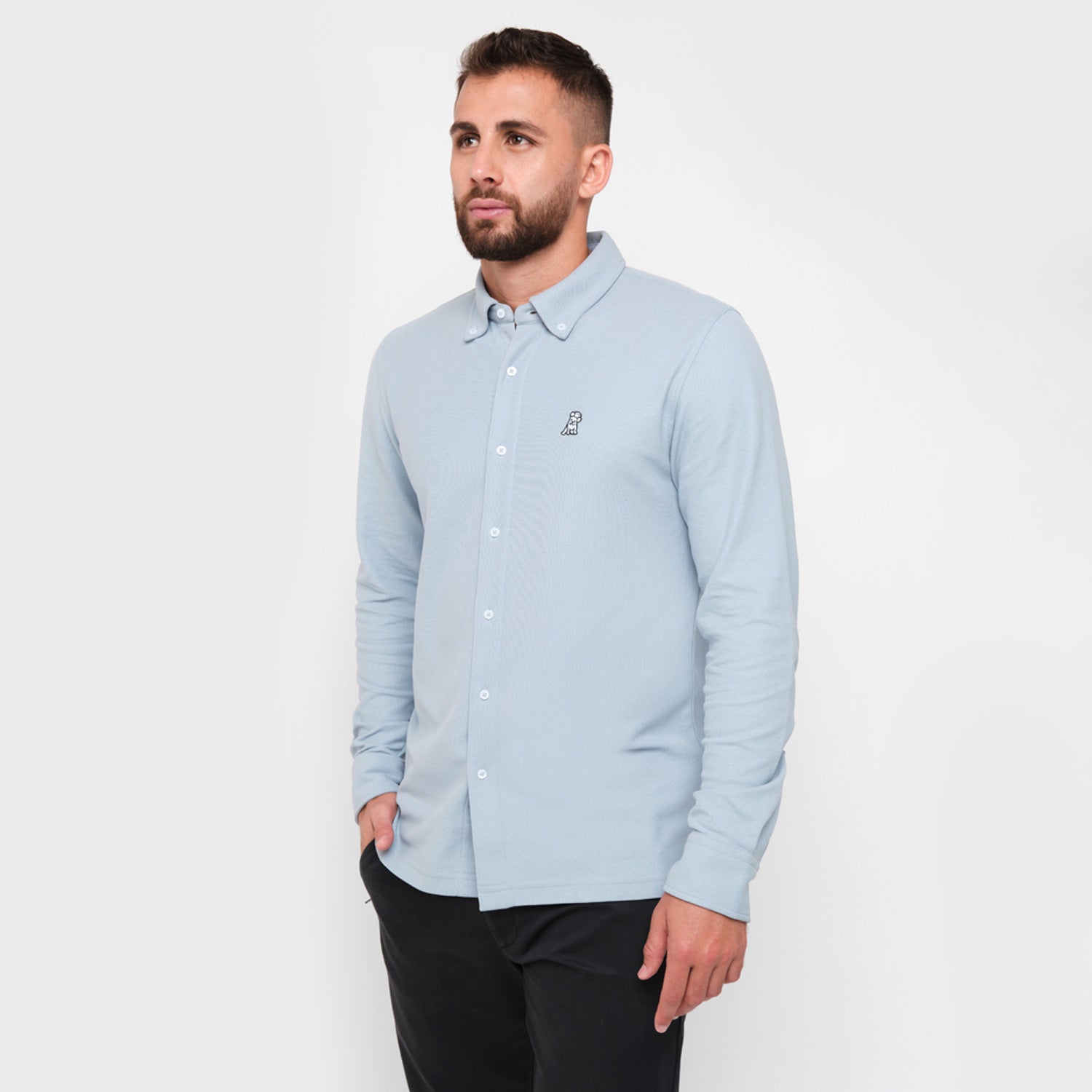 A man with a beard standing, wearing a light blue button-down shirt, looking to the side, paired with black pants.