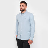A man with a beard standing, wearing a light blue button-down shirt, looking to the side, paired with black pants.