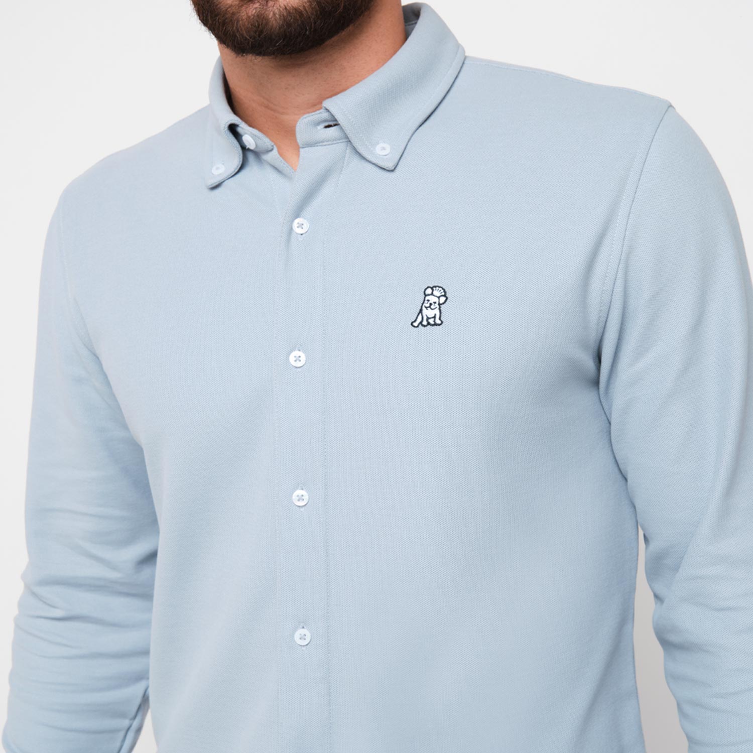 Close-up of a man wearing the light blue button-down shirt, showing the embroidered logo on the chest.