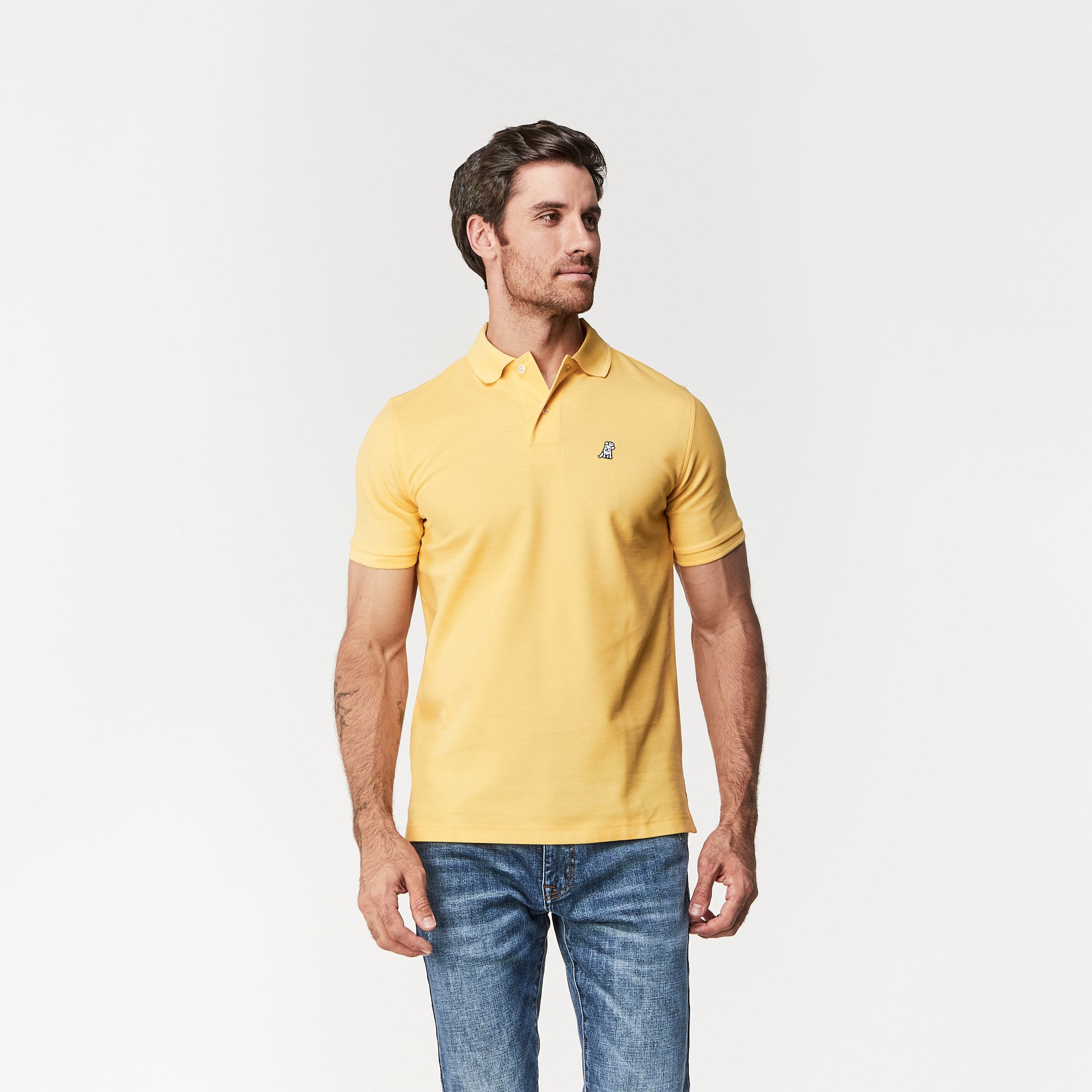 A man wearing a yellow polo shirt and jeans, standing in a casual pose, looking off to the side.