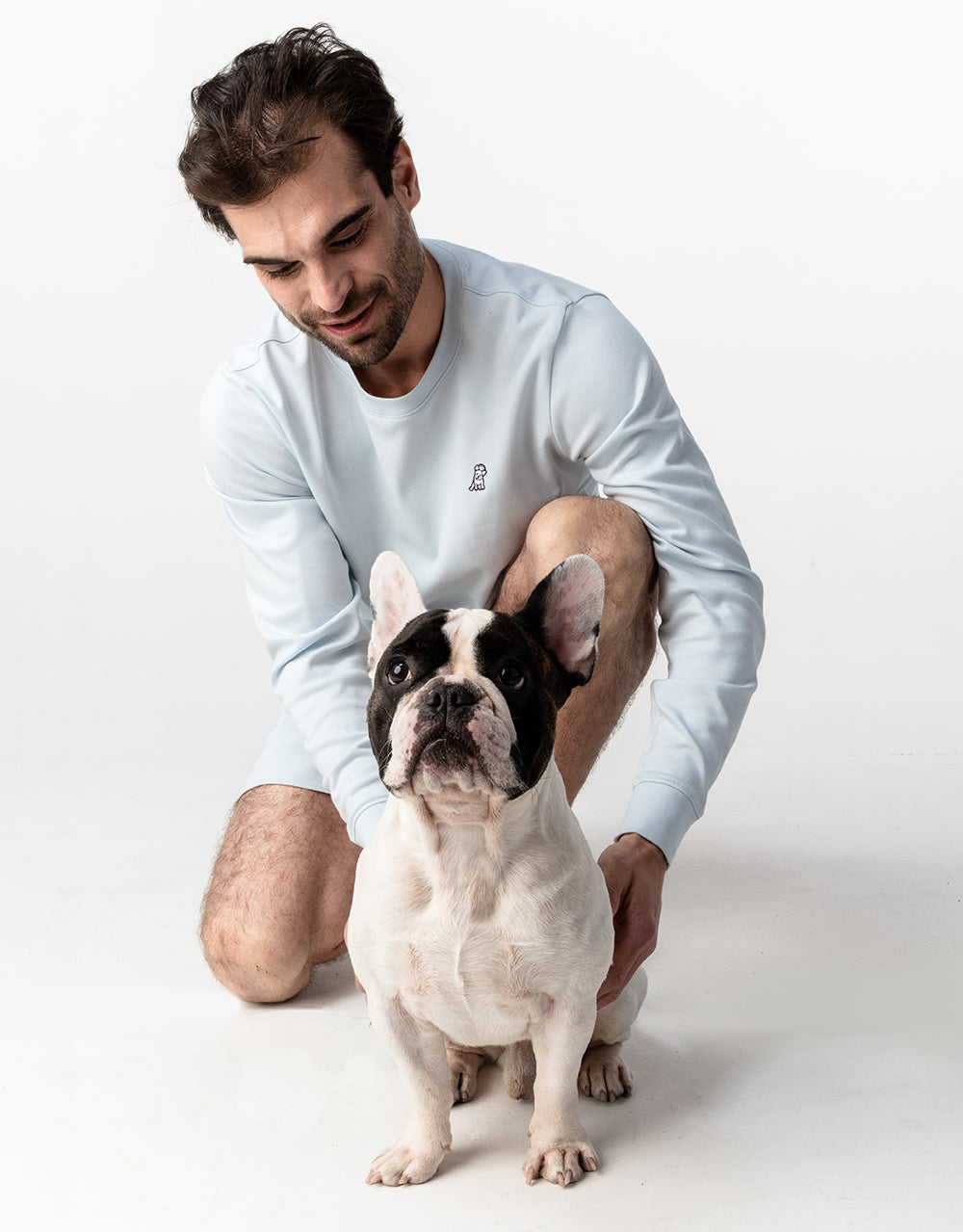 A man kneeling and smiling, looking at a black and white French Bulldog. The man is wearing a light blue long-sleeved shirt with a small embroidered dog logo