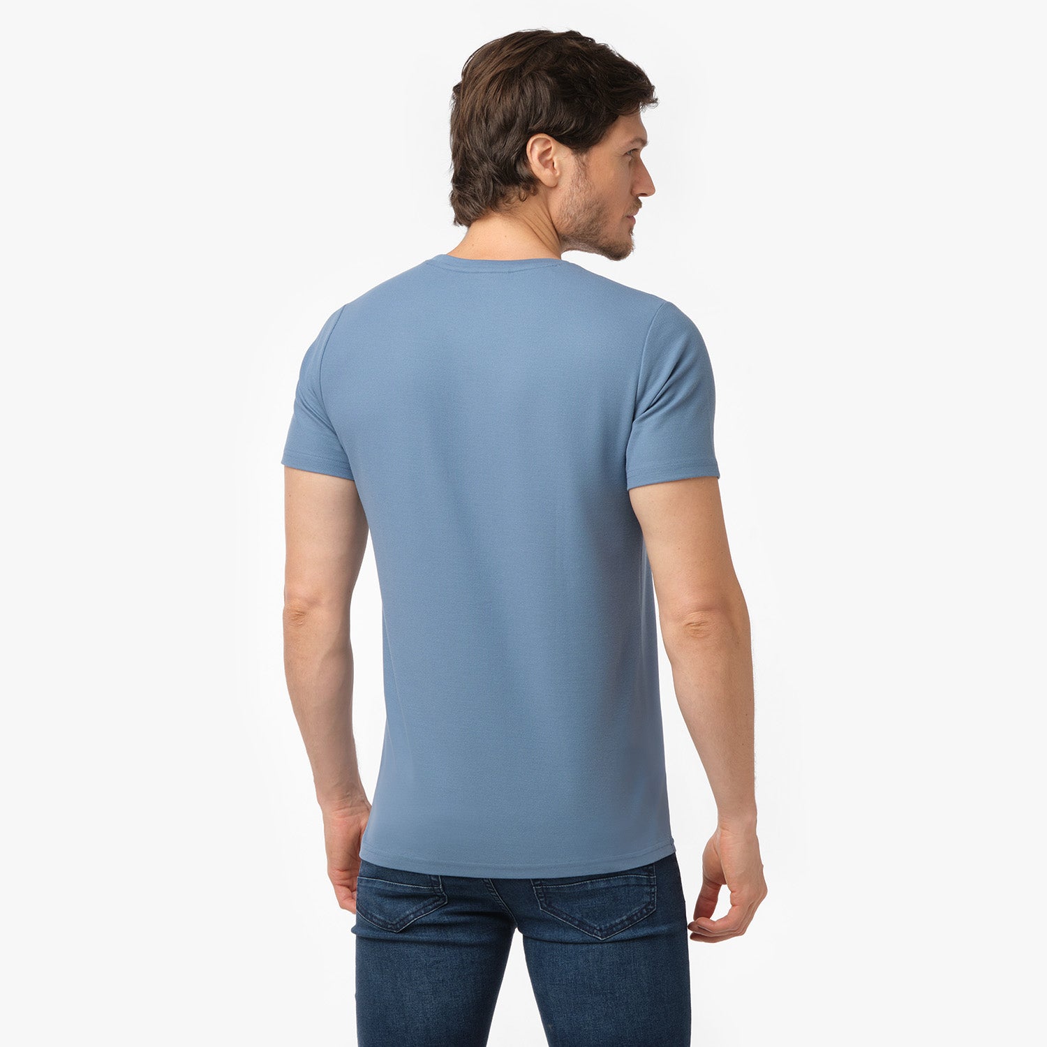 Male model showing the back of a light blue men's crew neck t-shirt and dark blue denim jeans, shot on white.