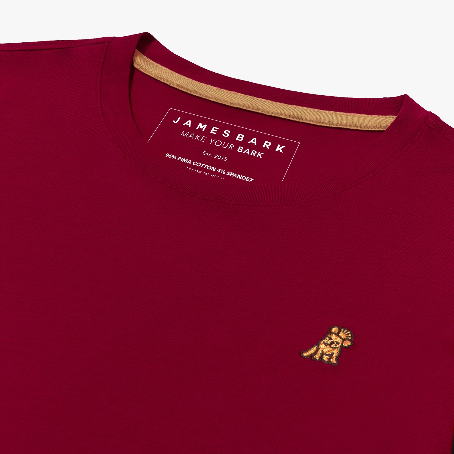 Close-up angled view of the inside collar area of the maroon crew neck t-shirt laid flat. Shows the ribbed neckline, gold neck tape, the printed "JAMES BARK" brand tag, and the gold/yellow embroidered dog logo on the chest