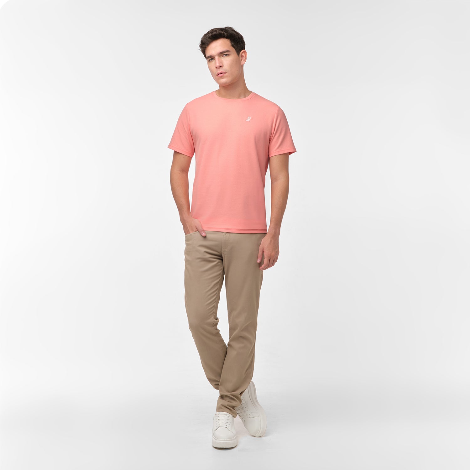 Full length photo of male model wearing a peach/coral men's t-shirt and tan chino pants, standing relaxed with one hand in his pocket, on white.
