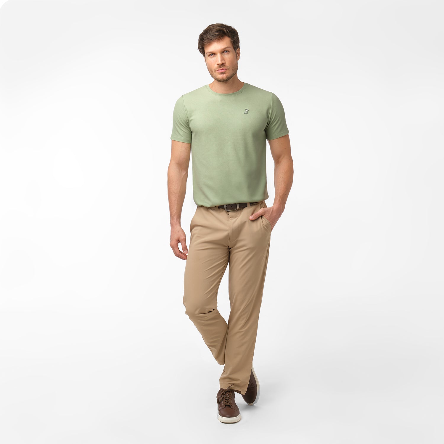Full length photo of male model wearing a light green men's t-shirt and tan chino pants, standing relaxed with one hand in his pocket, on white.