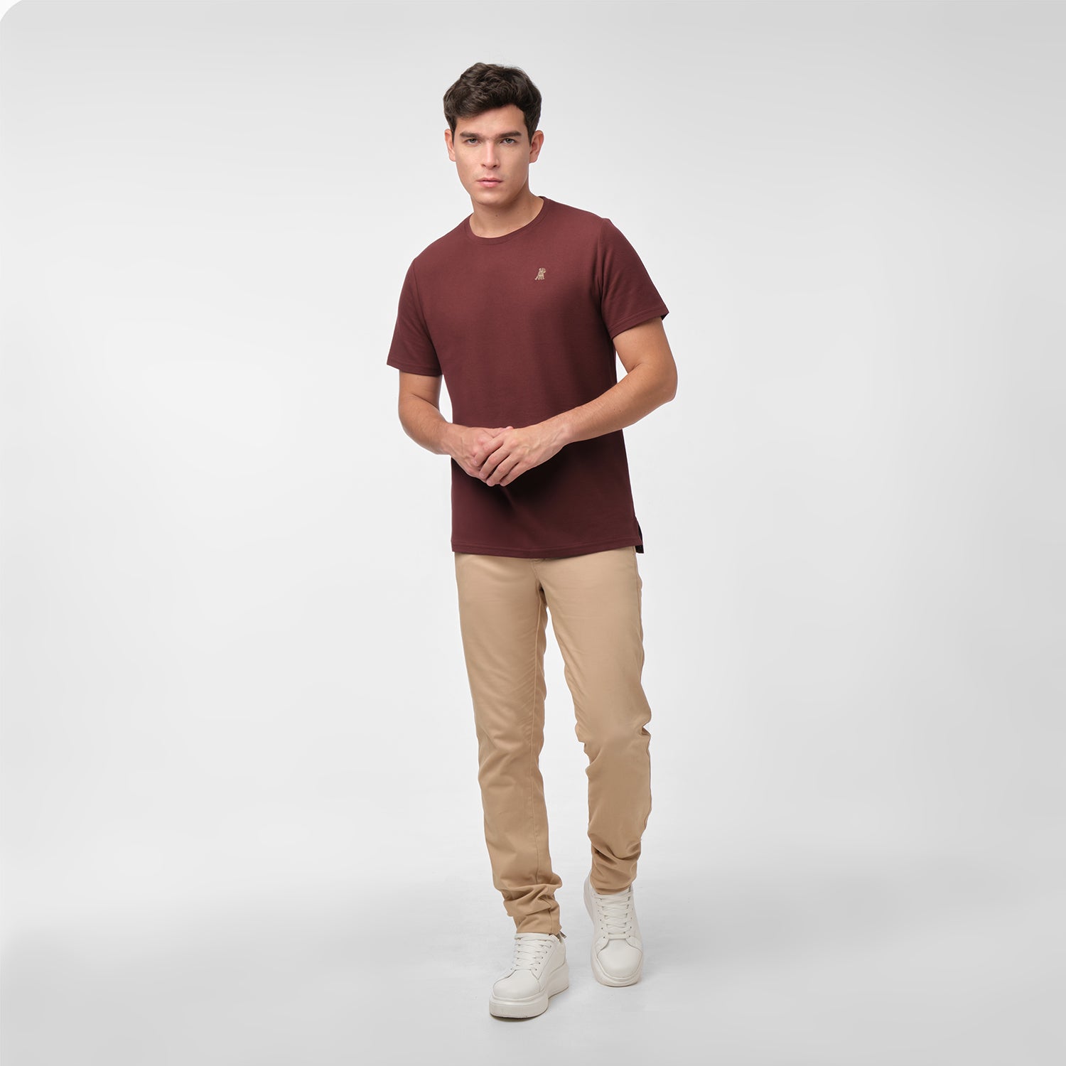 Full length photo of male model wearing a maroon men's t-shirt and tan chino pants, standing with hands clasped in front, on white.