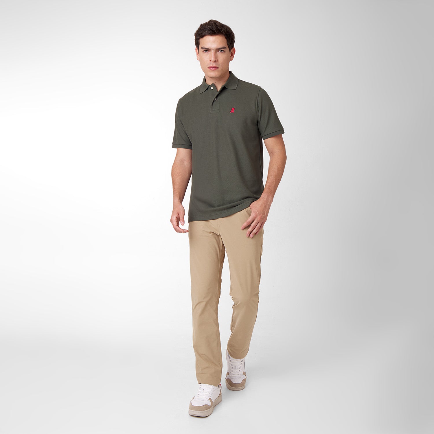 Full body picture of a male model wearing an olive green polo shirt with a red embroidered dog logo and tan pants, standing front view