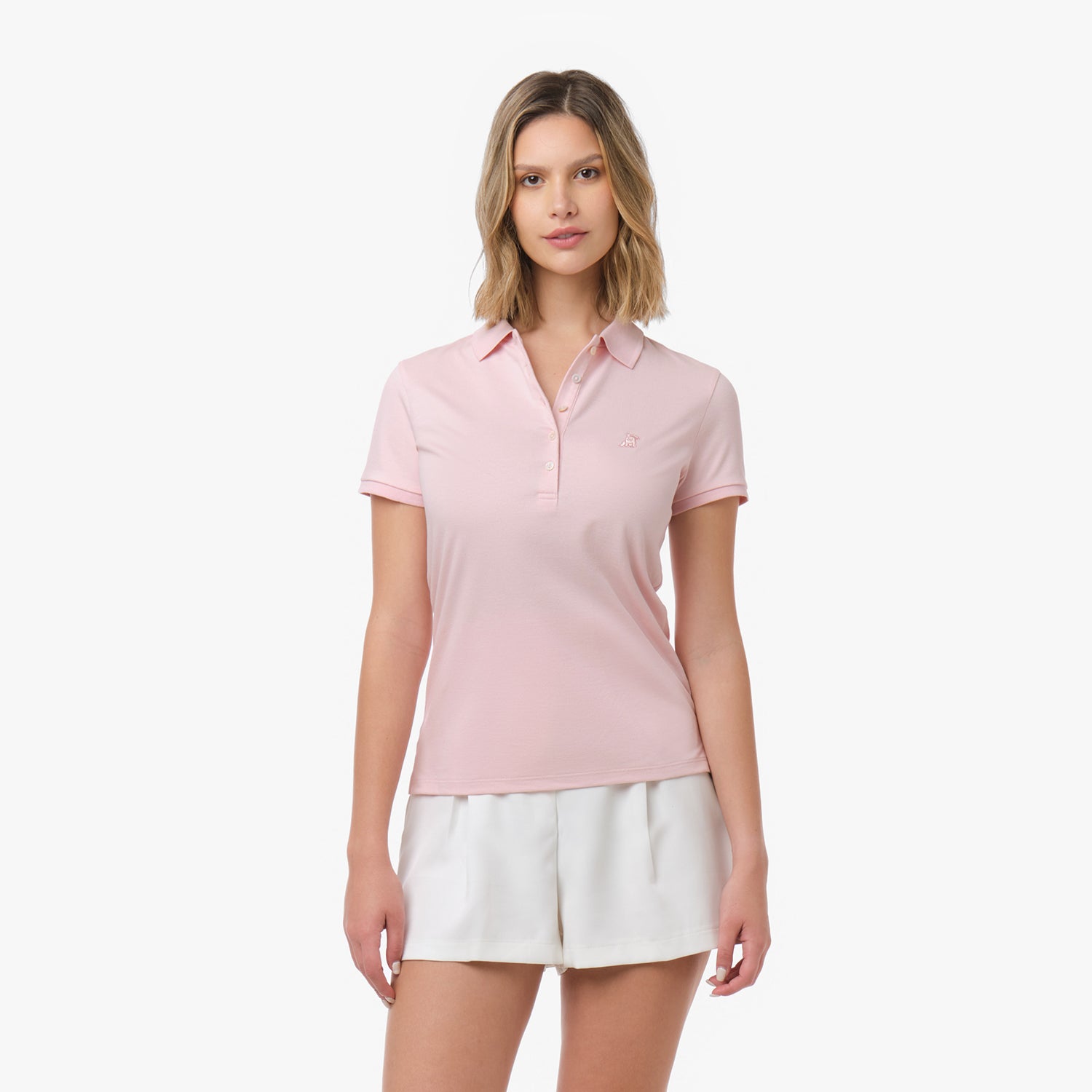 Woman models a light pink short-sleeve polo shirt with white buttons and tonal embroidered logo, paired with white shorts. Front view against white background