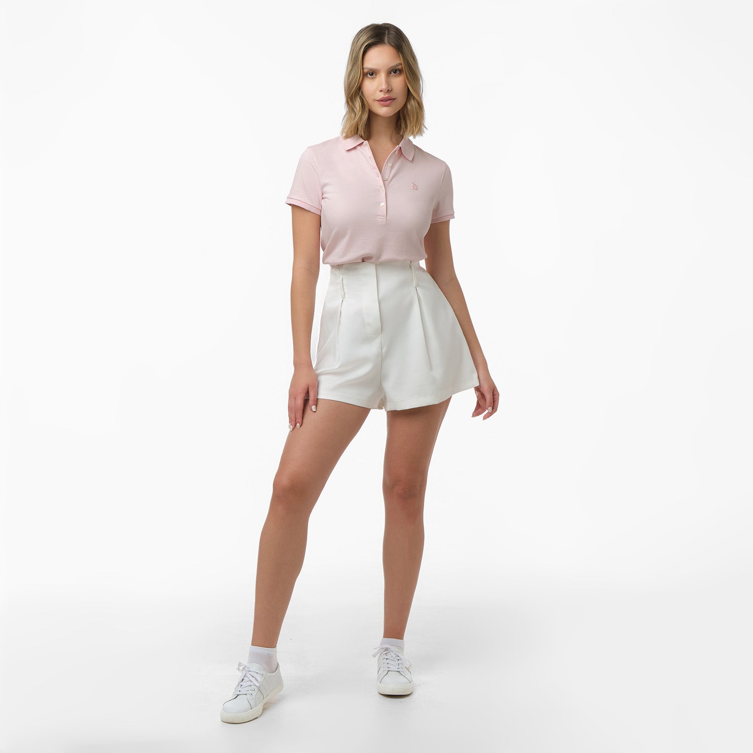 Full-length view of a woman modeling a light pink polo shirt, high-waisted white shorts, white socks, and white sneakers. Standing pose against white background