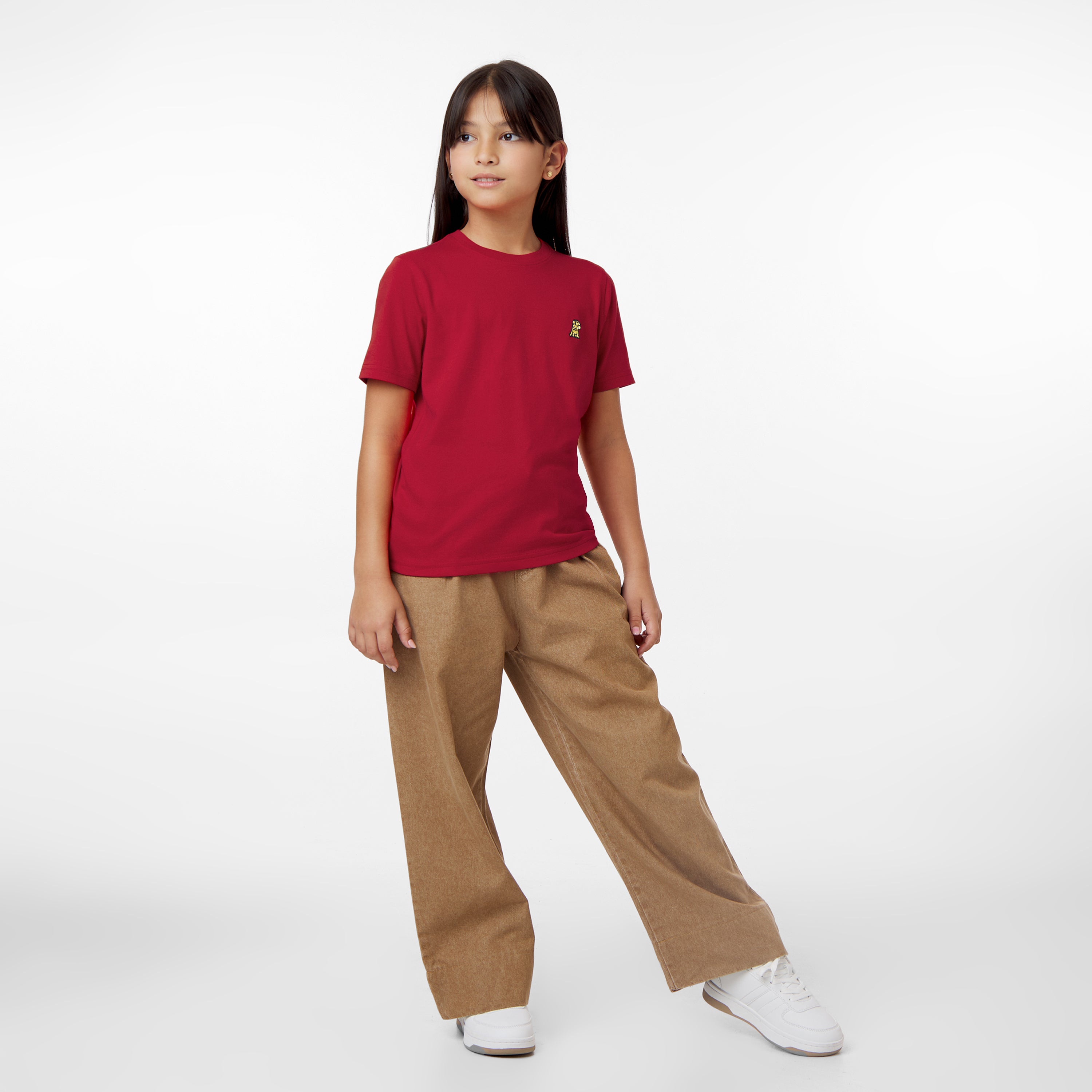 A young girl stands posing in a red t-shirt with a small logo and brown pants, looking to the side, against a white background.