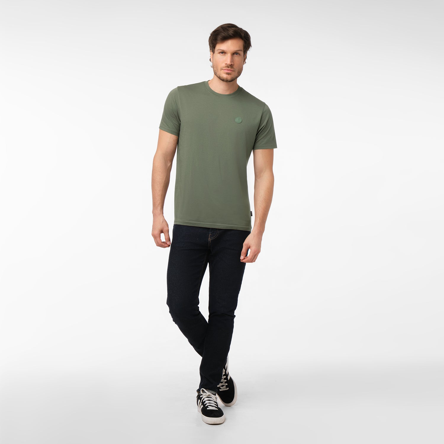 Full-length view of a male model wearing an olive green crew neck t-shirt with a tonal embossed circular logo, dark blue jeans, and black sneakers, standing with legs crossed