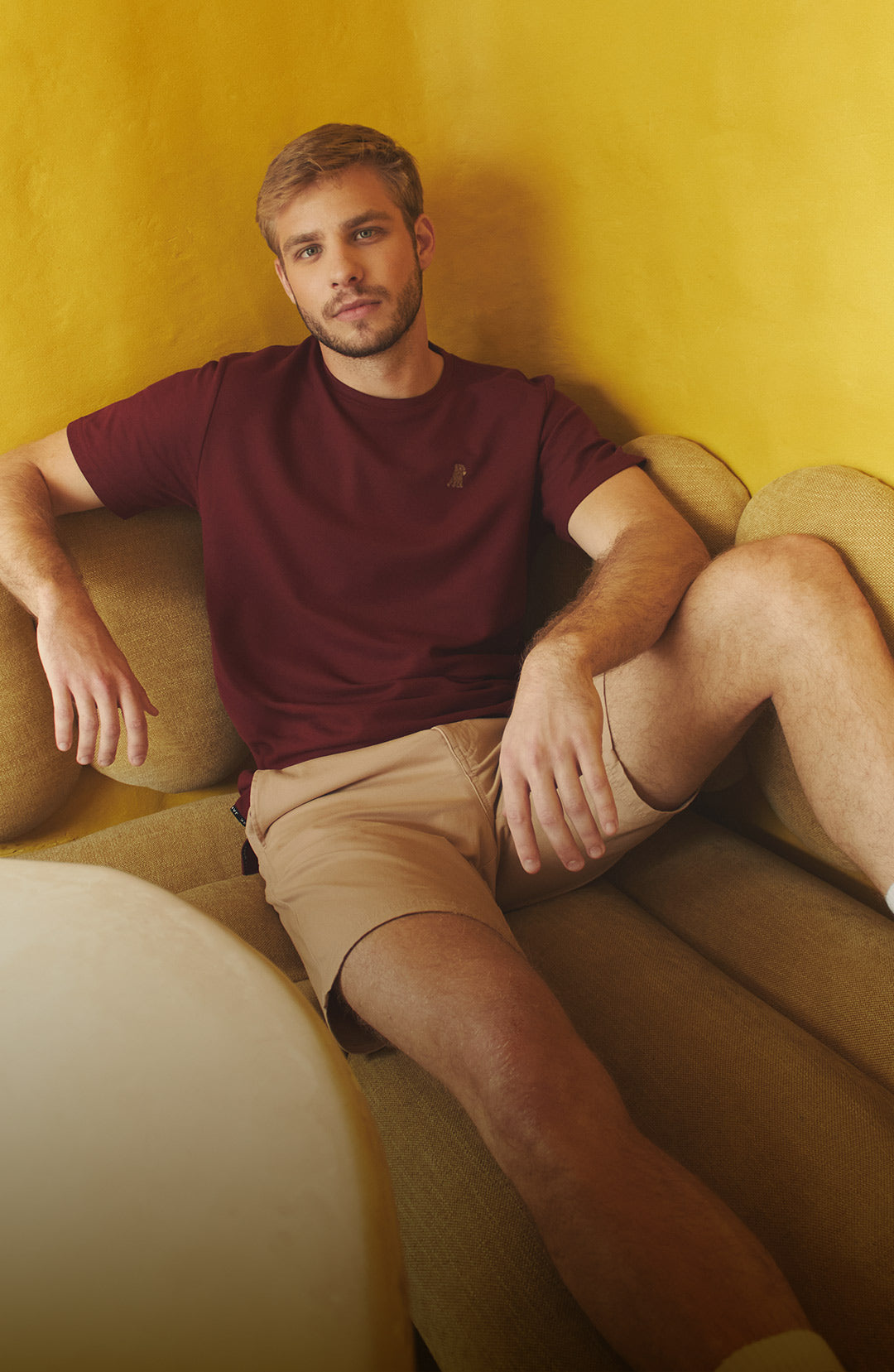 A man sits on a textured, curved couch in front of a yellow wall, wearing a maroon t-shirt, khaki shorts, and white socks with black stripes. He is looking at the camera. A blurry cream table is in the foreground.