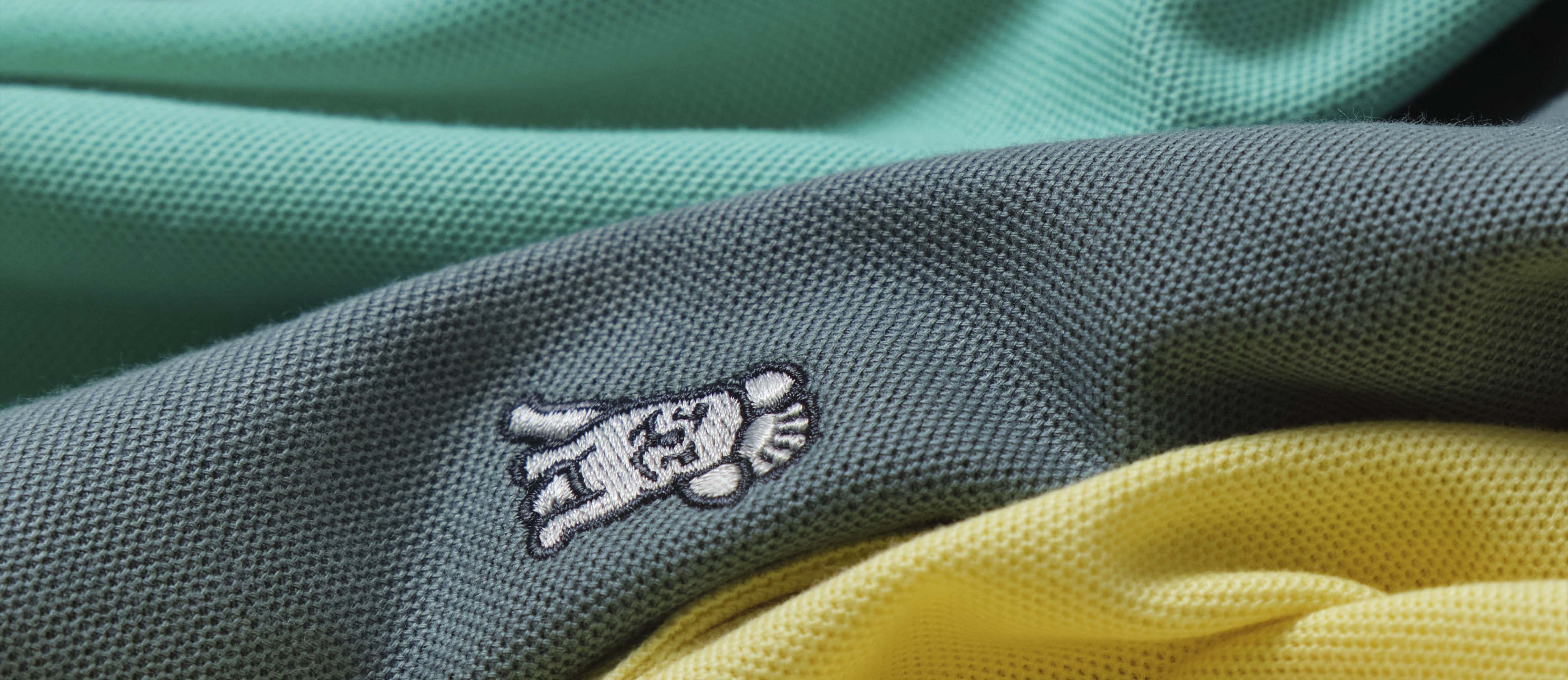 Close-up of colorful fabric in shades, each featuring an embroidered logo of a cartoon-style dog. The fabrics are laid on top of each other, highlighting the texture and detail of the embroidery.