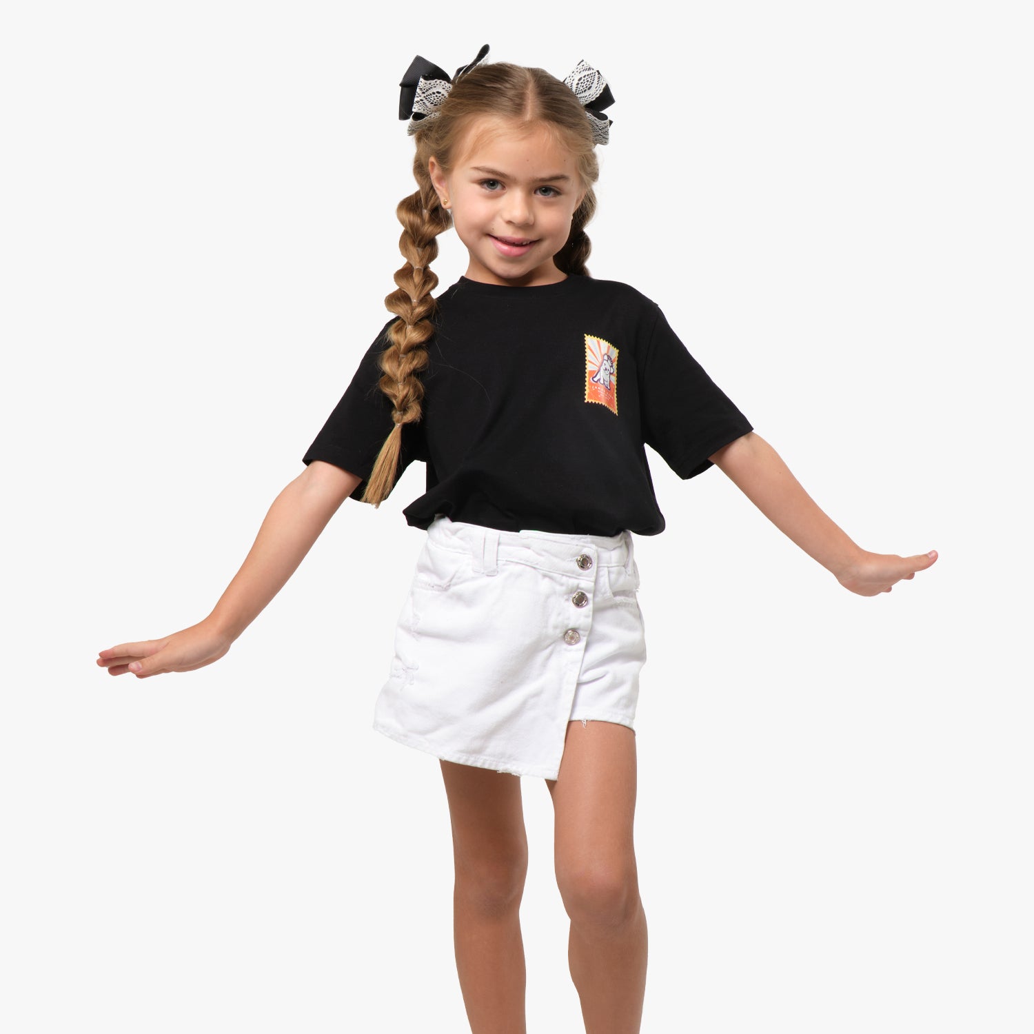 A young girl wearing a black T-shirt with a colorful graphic on the chest, paired with a white skirt. She is playfully posing with her arms outstretched.