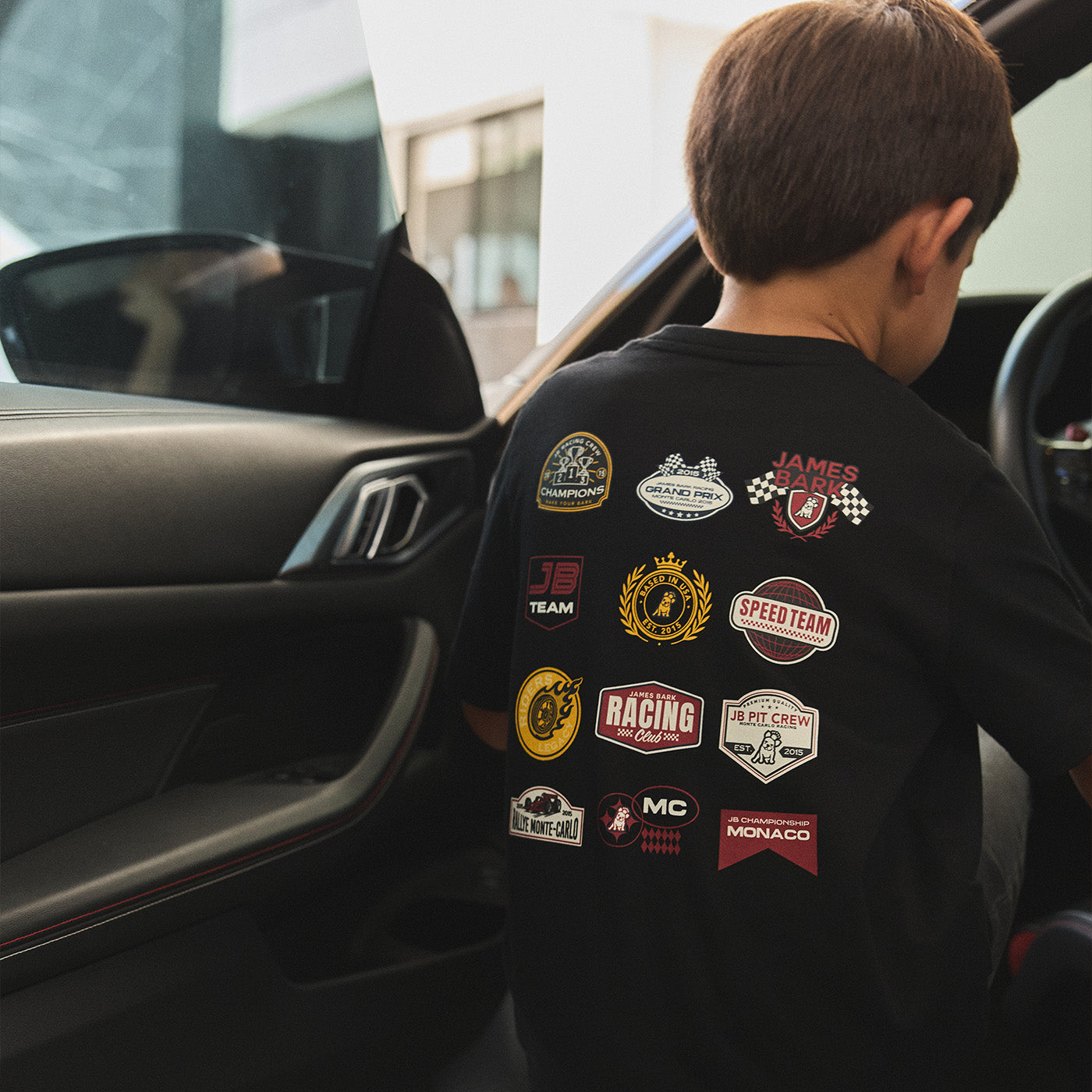 Young boy sitting in a car wearing a black James Bark racing-themed graphic t-shirt with multiple motorsport logos.