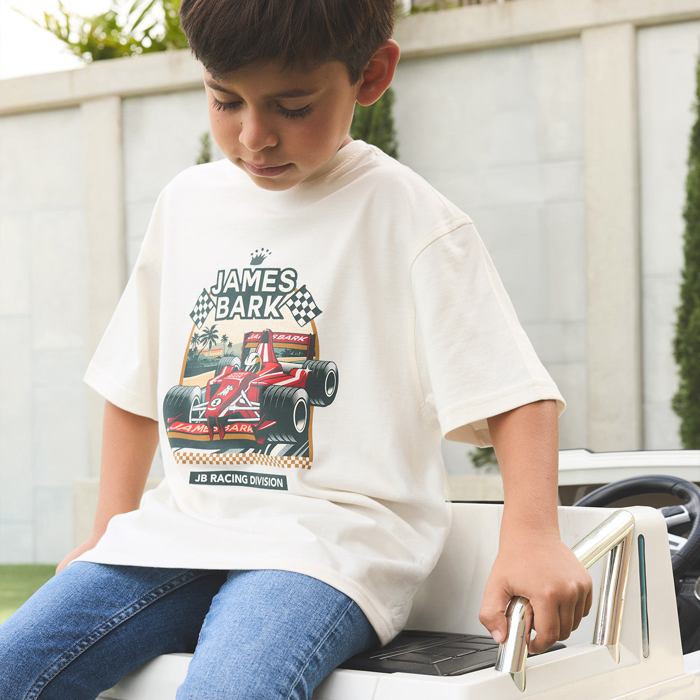 Young boy wearing a cream James Bark 'Racing Division' graphic t-shirt with a vintage race car print.