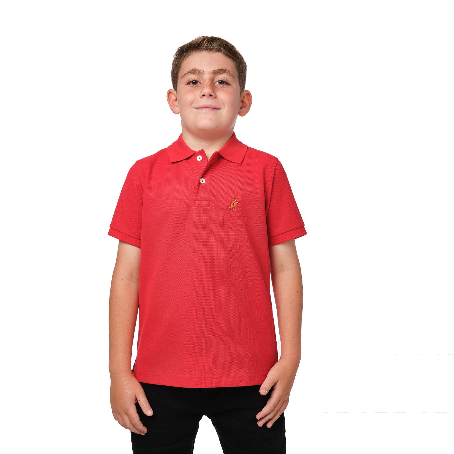 A young boy wearing a red polo shirt with a small gold embroidered logo on the chest, standing with his hands at his sides and smiling.