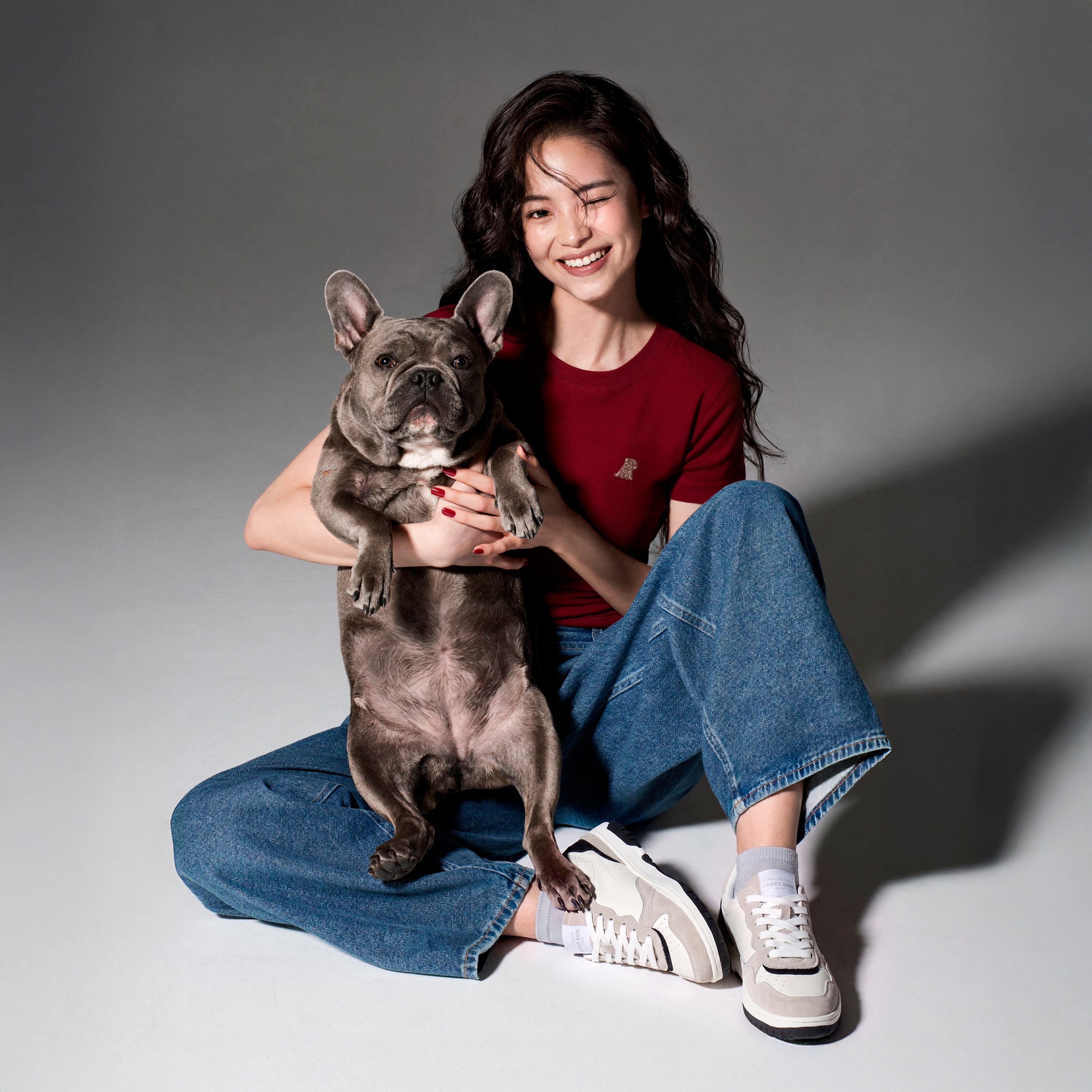 A woman sitting on the floor holds a gray French bulldog. She is wearing a dark red short-sleeve t-shirt with a small white logo on the chest, blue jeans, and white sneakers.