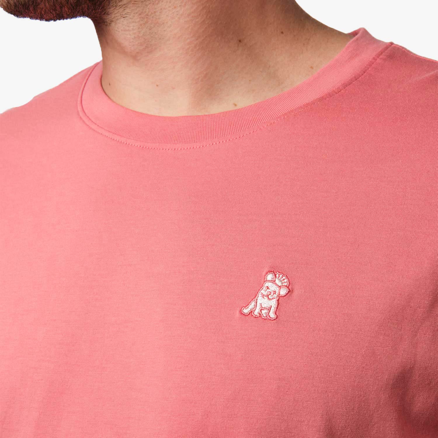 Close-up of the upper chest area of the salmon pink t-shirt, focusing on the small embroidered animal logo near the neckline.