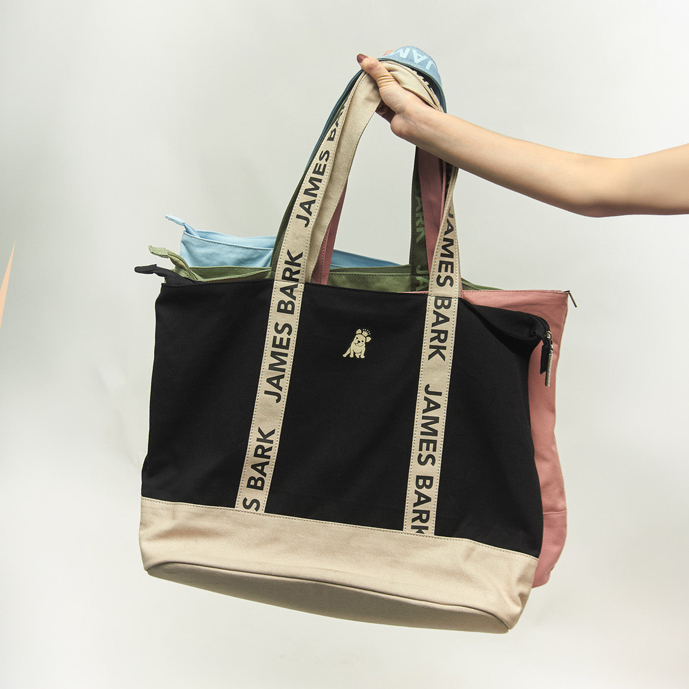 Hand holding three James Bark canvas tote bags in black, green, and light blue with branded shoulder straps