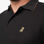 Close-up of the embroidered logo on the black polo shirt: A gold embroidered dog logo is visible on the left chest area, adding a subtle detail to the polo.