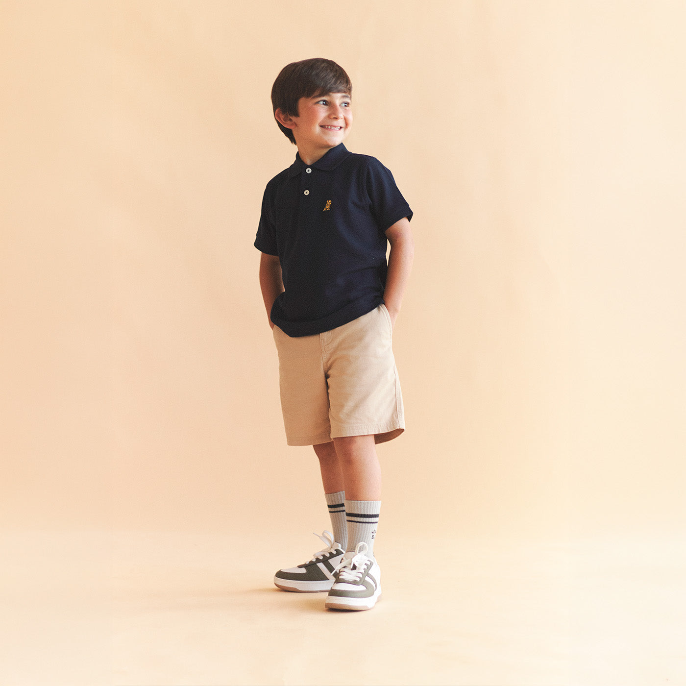 Boy wearing a navy polo with khaki bermuda shorts on a light beige background