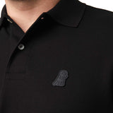 Close-up of the black embroidered logo on the chest of the black polo shirt.
