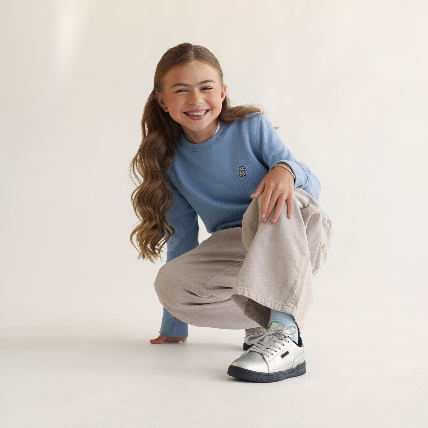 Young girl wearing a light blue James Bark sweatshirt and beige wide-leg pants, crouching down while wearing silver sneakers.
