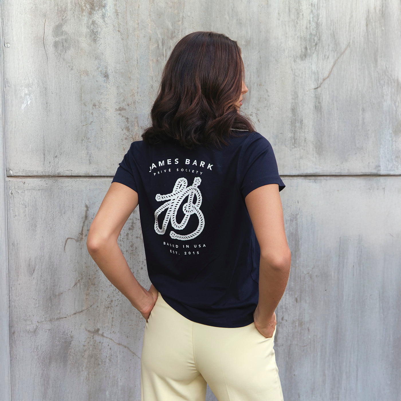 Woman wearing a navy blue t-shirt with a logo and text against a concrete wall.