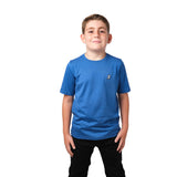 Front view of a boy wearing a blue t-shirt with a small white embroidered logo on the chest, standing with his hands in his pockets.