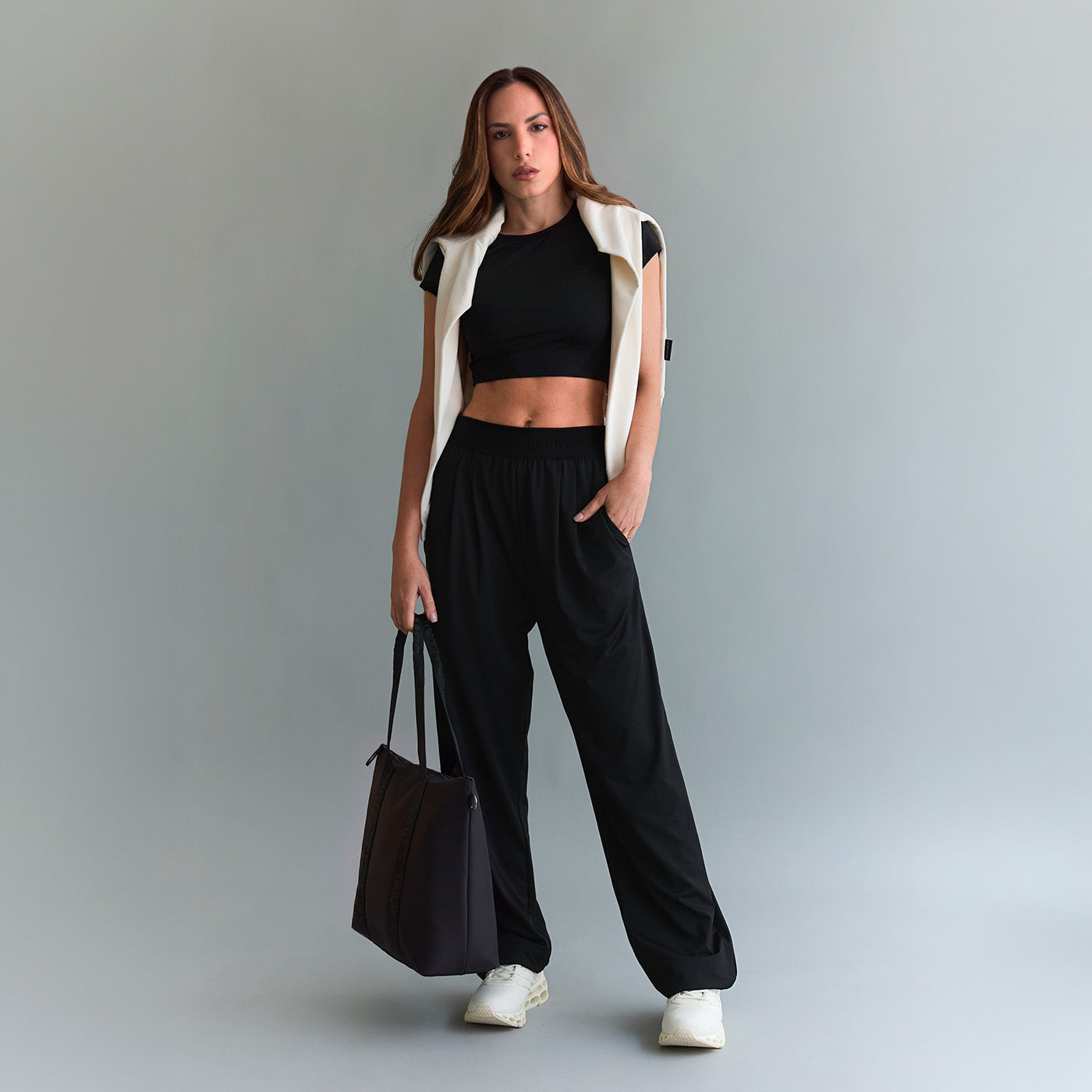 Woman wearing a black cropped t-shirt and black joggers, styled with a white sweatshirt draped over shoulders and a black tote bag.
