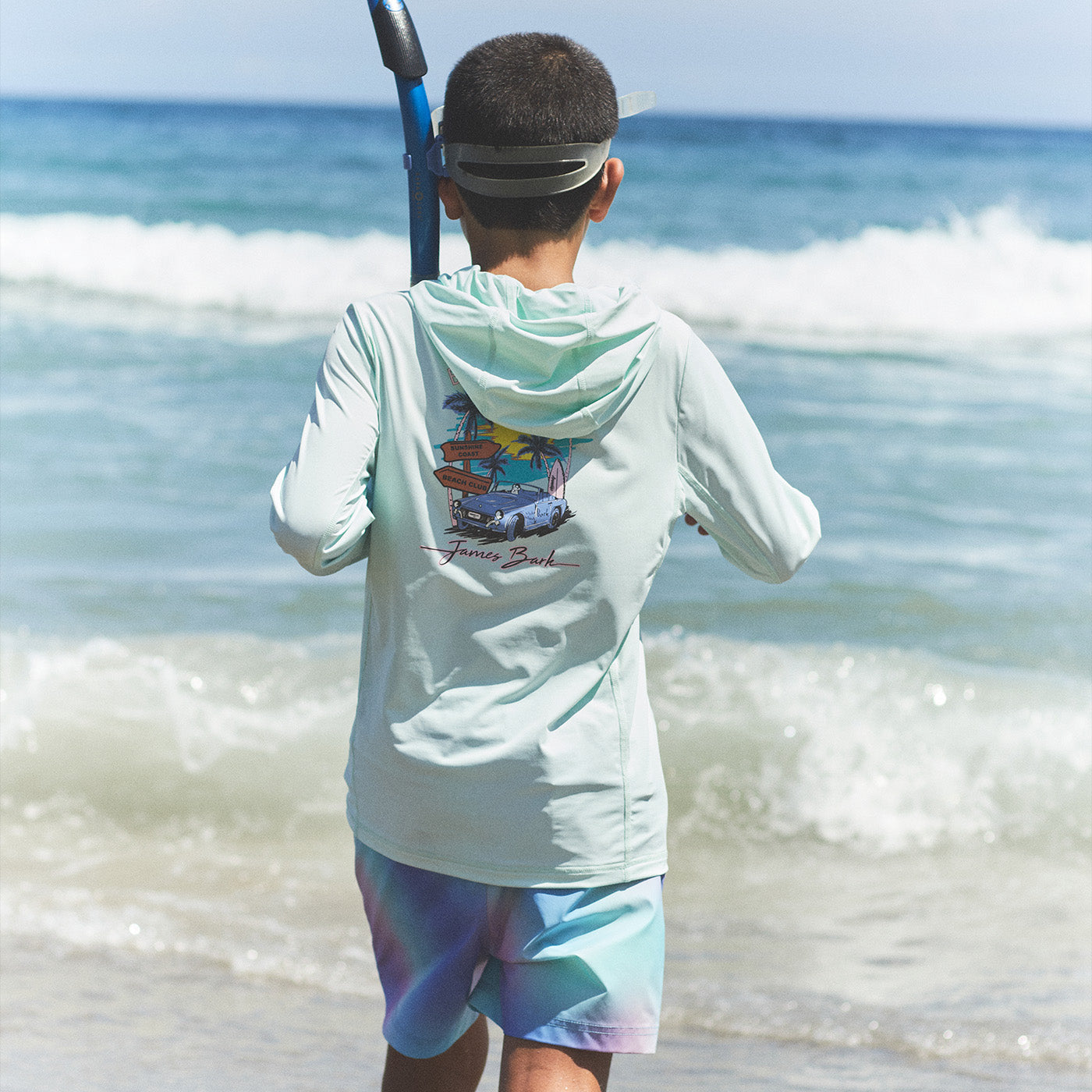 Young boy wearing a mint green James Bark hooded rashguard with a graphic print, standing on the beach with snorkel gear.