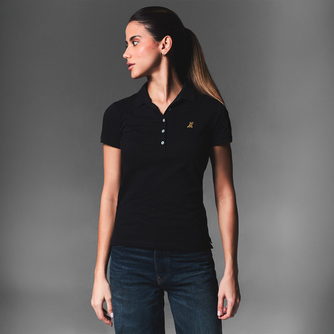 Woman wearing a black James Bark polo shirt and dark blue jeans, standing against a grey background.