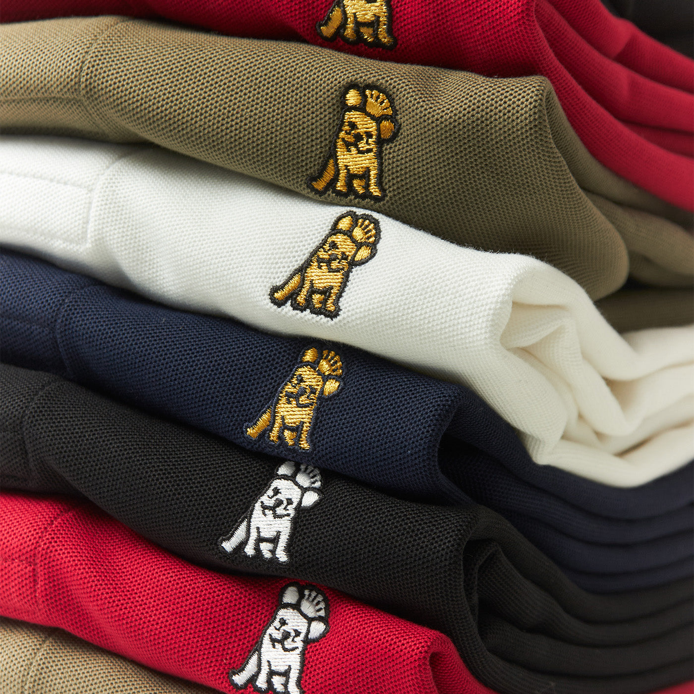 Stack of folded James Bark polo shirts in various colors, showcasing embroidered Frenchie logo detail.