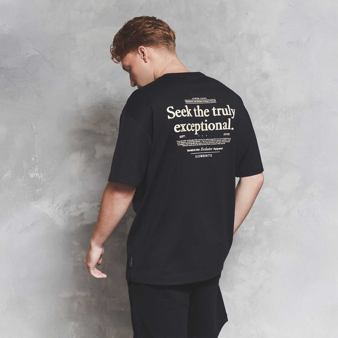 Person wearing a black t-shirt with text on the back against a gray concrete wall