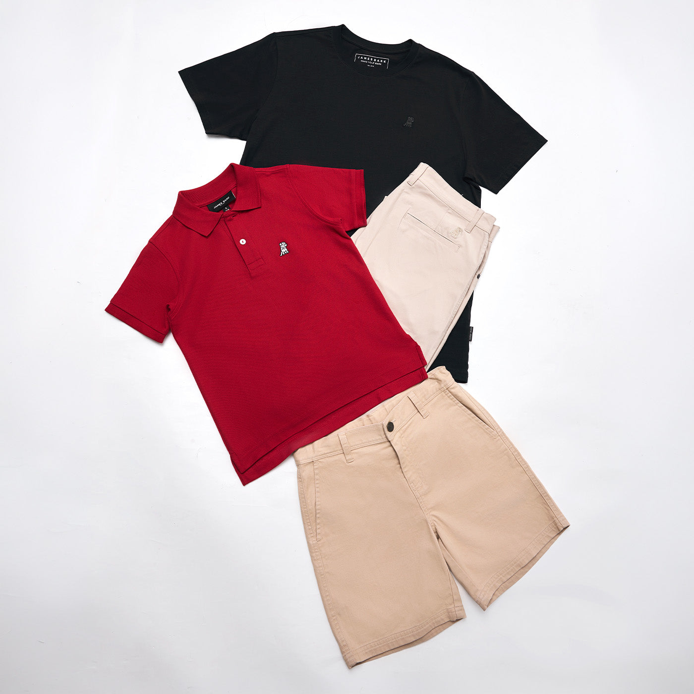 Flat lay of James Bark apparel on a white background, featuring a black crew neck T-shirt, a red pique polo shirt with the signature logo, and beige chino shorts
