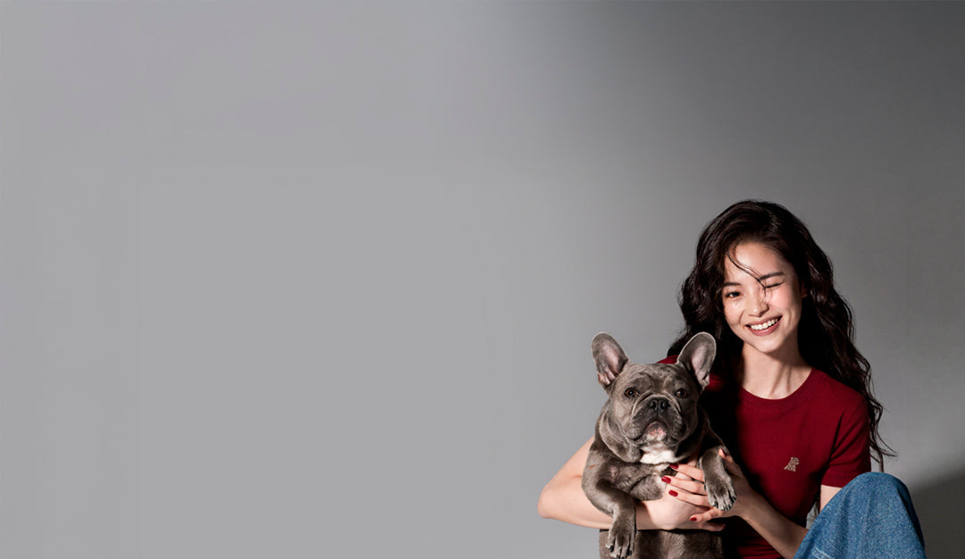 A woman with long dark hair smiling while holding a gray French bulldog, wearing a red t-shirt and blue jeans, set against a solid gray studio background.