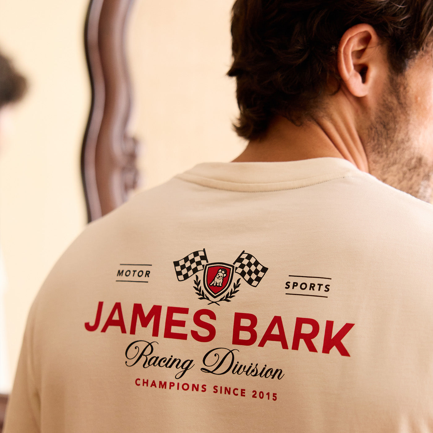 Man wearing a cream James Bark Racing Division graphic t-shirt, viewed from behind.