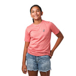 Front view of a girl wearing a pink t-shirt with a small white embroidered logo on the chest, smiling with one hand in her pocket.