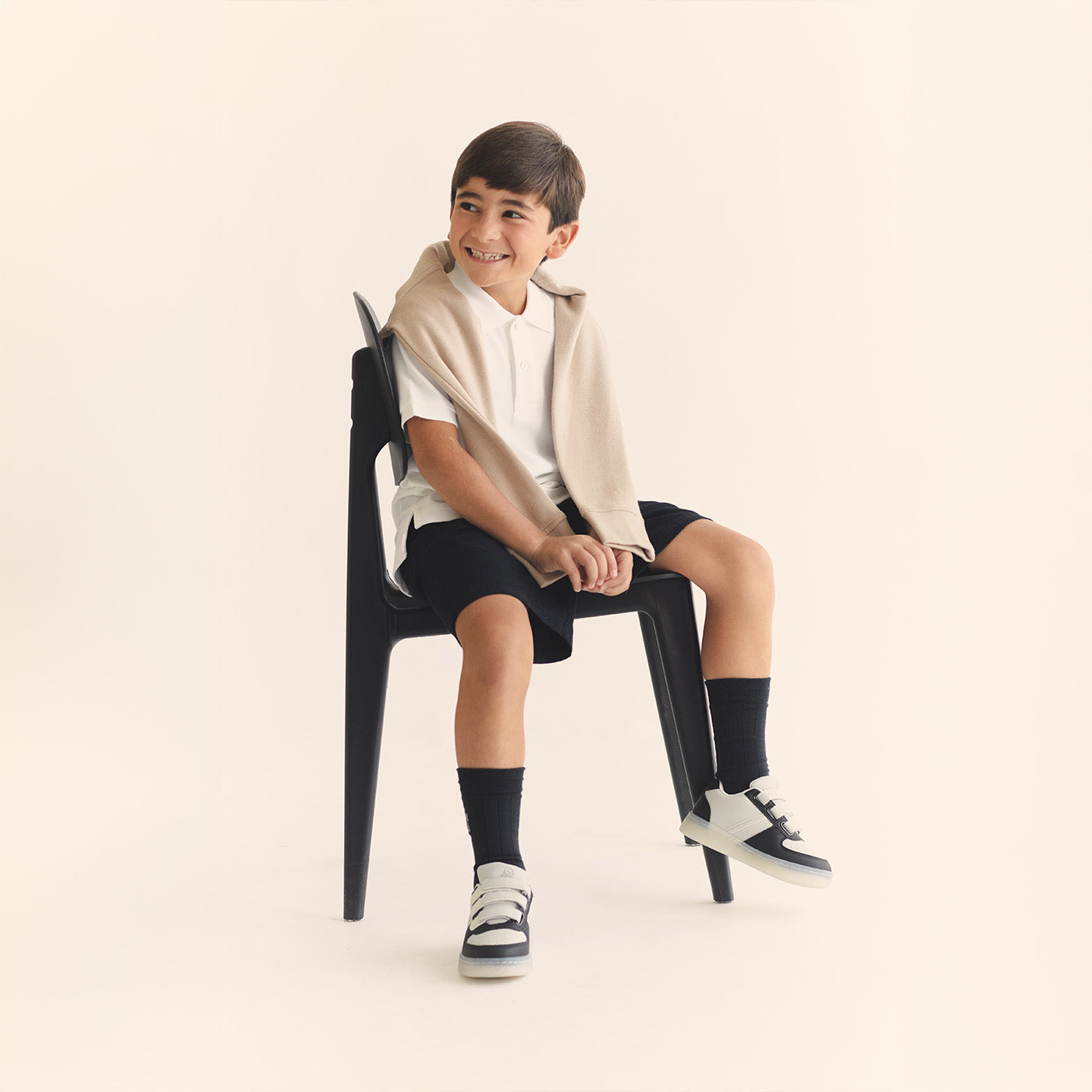 Child sitting on a black chair wearing a beige sweater, white shirt, black shorts, and black socks with white shoes.