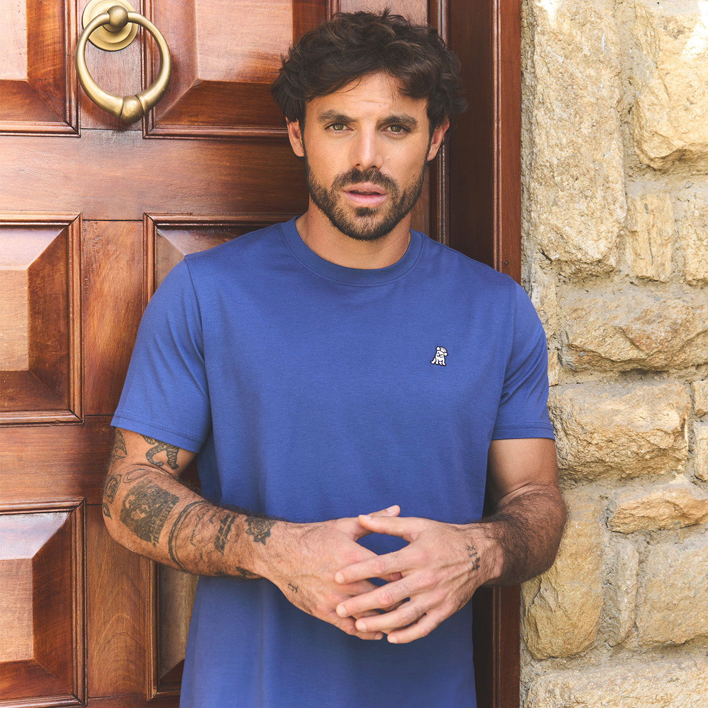 Man wearing a blue t-shirt with a small logo, standing in front of a wooden door 