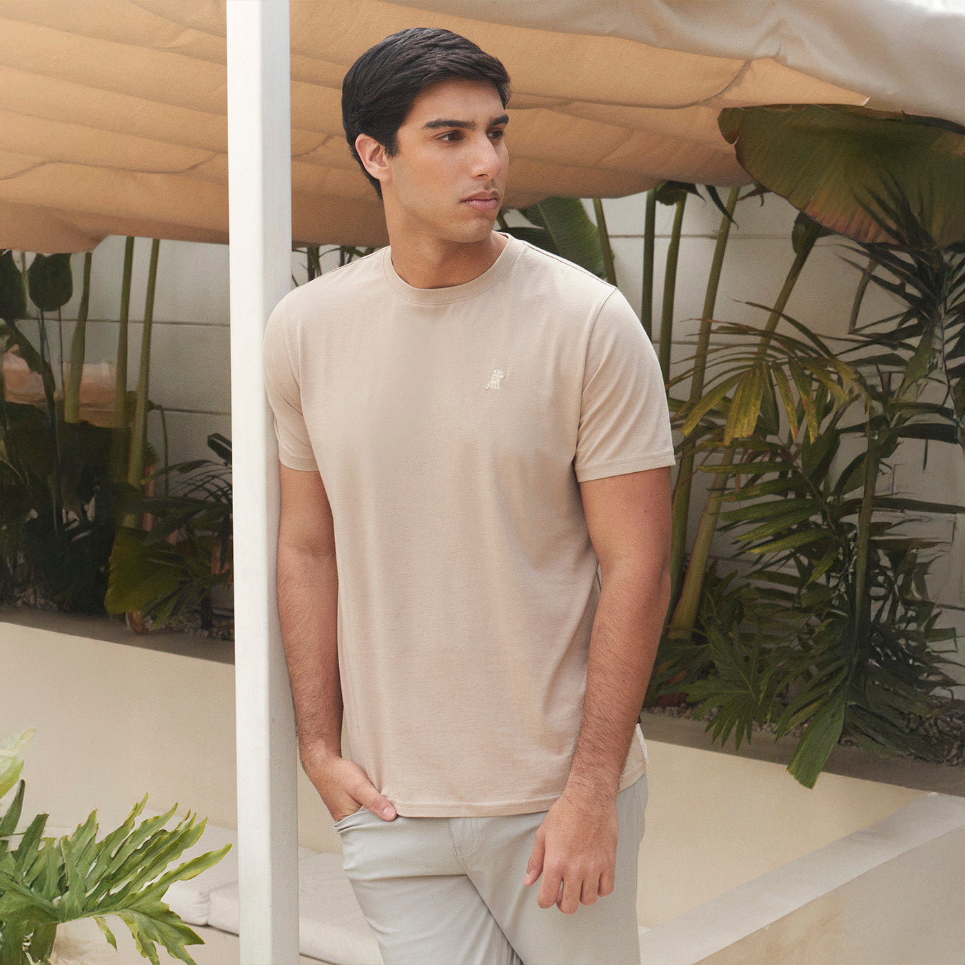 Man wearing a beige t-shirt with a logo, standing in an indoor setting with plants.
