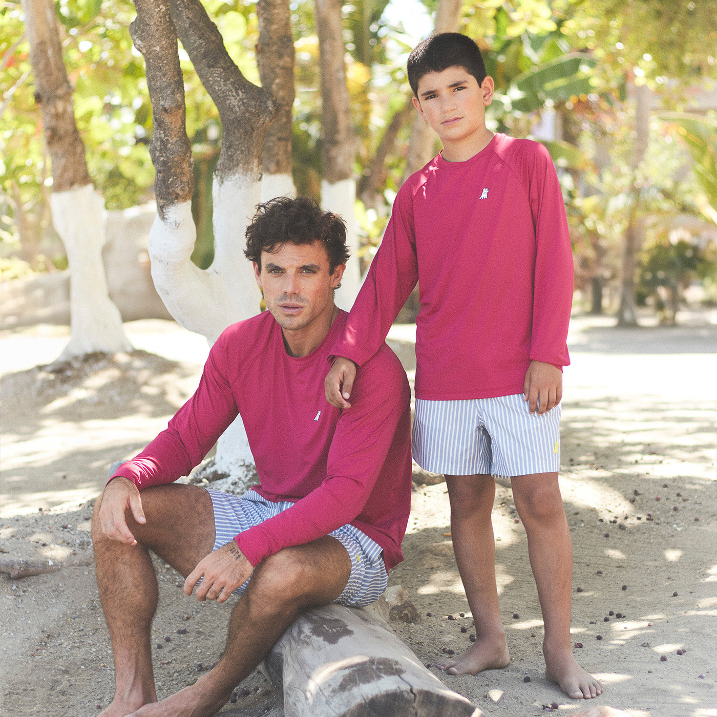 Matching James Bark Men's and Kids' Beachwear: Father and son in magenta long-sleeve rashguards and blue striped swim trunks