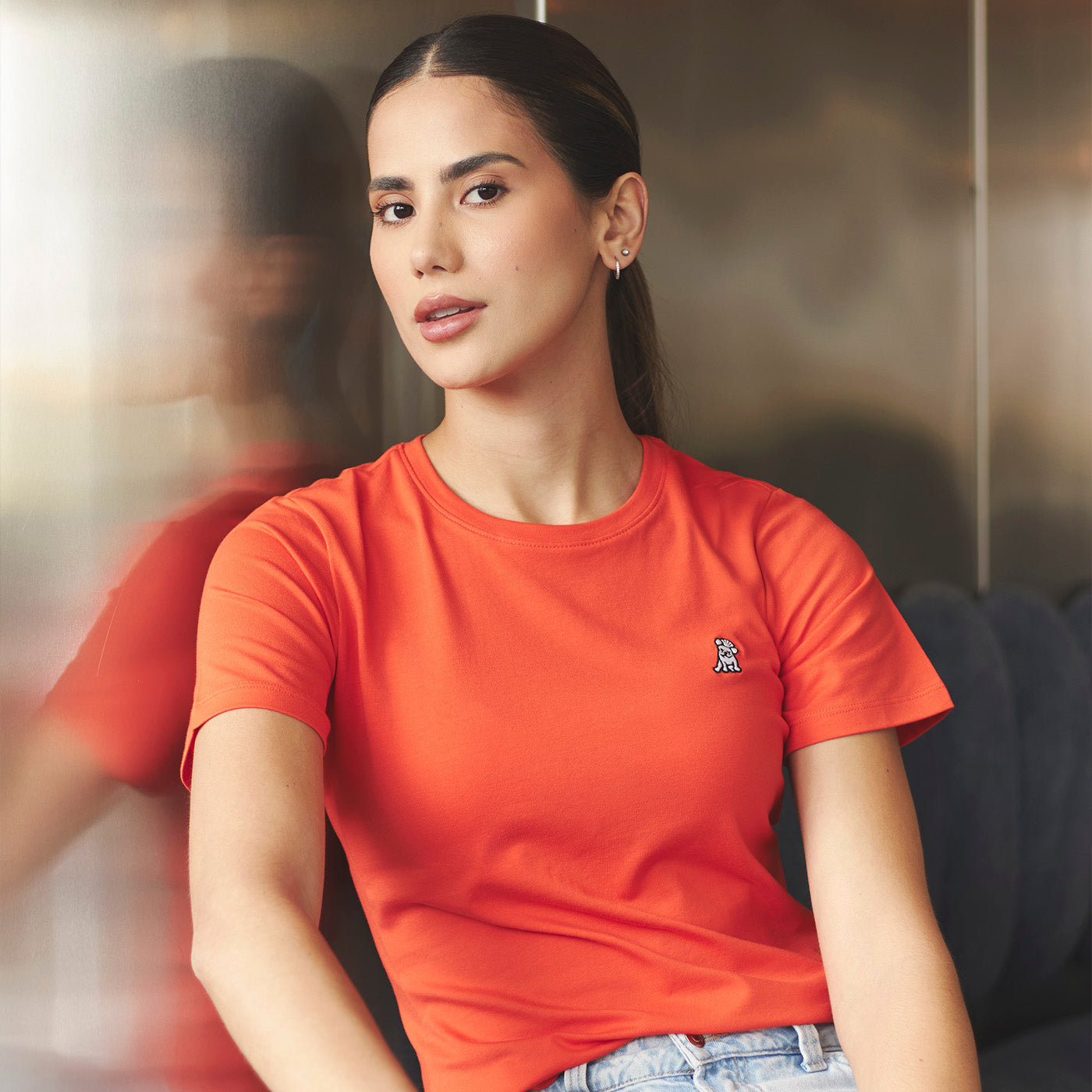 Woman wearing an orange t-shirt with a small logo, sitting indoors.