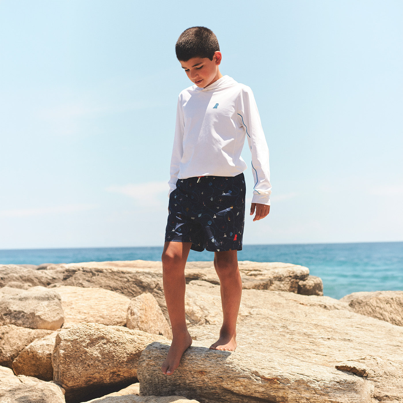 Child standing on rocks by the ocean wearing a white long-sleeve shirt and dark shorts.