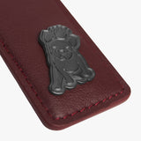  A close-up view of a burgundy leather tag with a metallic emblem of a dog wearing a crown, highlighted by contrasting red stitching.