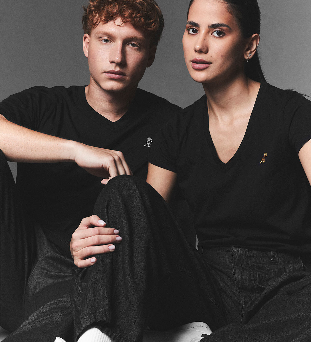 Editorial shot of a couple sitting on the floor in black v-neck t-shirts—one with a white logo, the other with a gold logo—styled with black pants and white sneakers.