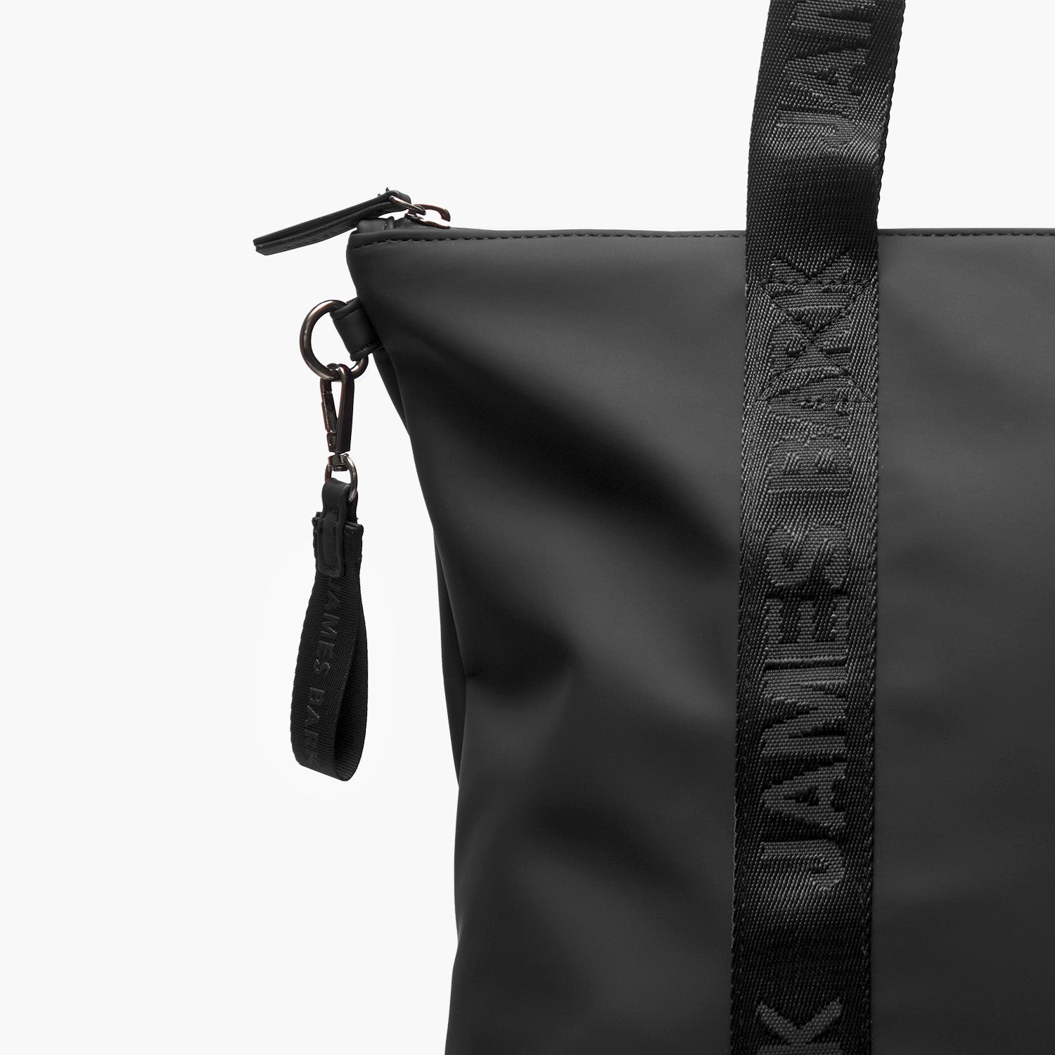 A close-up of a black tote bag highlighting its details. The bag features a matte finish, a sturdy zipper closure at the top, and a detachable wrist strap with the brand name "JAMES BARK" embossed on it. The bag's handles are made of thick, durable fabric with the same embossed branding running along their length. The stitching and metal hardware, including a clasp and ring for additional attachments, add to the bag's premium and functional design.